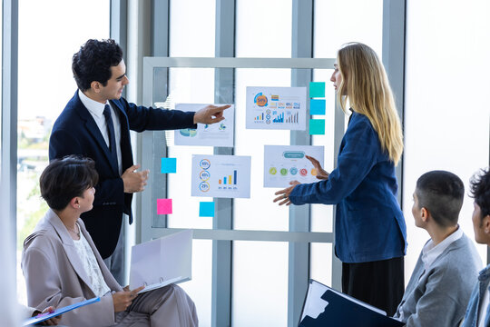 Professional businesswoman writing ideas on a glass presentation board with a marker while explaining financial data to a thoughtful male colleague in a modern office.