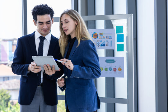 Professional businesswoman and businessman discussing data on a digital tablet near a glass presentation board in a modern office environment.