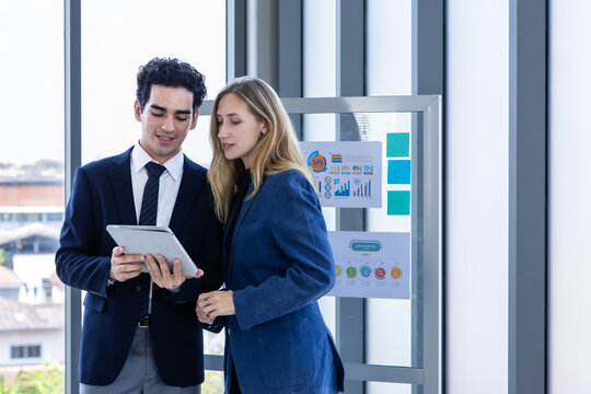 Professional businesswoman and businessman discussing data on a digital tablet near a glass presentation board in a modern office environment.