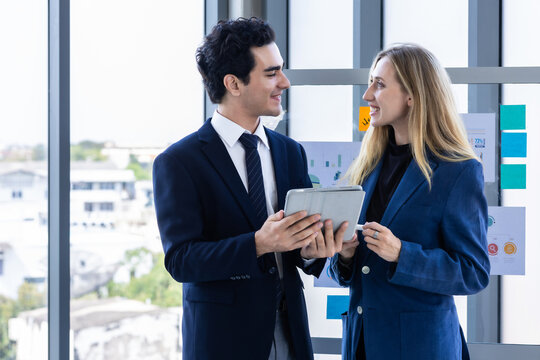 Professional businesswoman and businessman discussing data on a digital tablet near a glass presentation board in a modern office environment.