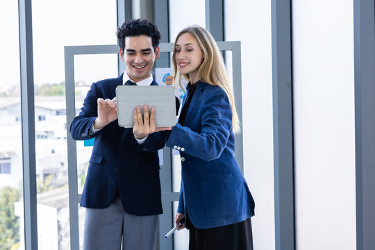 Professional businesswoman and businessman discussing data on a digital tablet near a glass presentation board in a modern office environment.