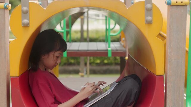 Asian kid drawing digital art on tablet inside a playground tunnel. Selective focus highlights the creative elementary student enjoying casual hobby and relaxing time outdoors with technology.