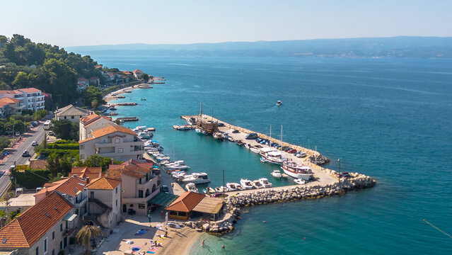 High-angle drone shot showing a scenic coastal village with red-roofed houses, a busy harbor with various boats, and a stone pier along the clear blue water.