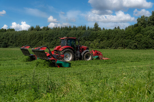 Red tractor mowing a green forage field beside a summer forest
