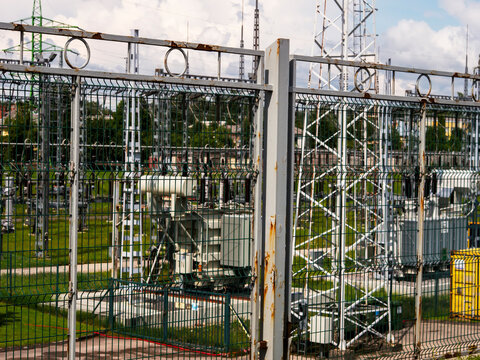 Metal fence surrounds industrial electrical substation. Transformers and towers visible behind fence. Green grass and trees around facility. Sky partly cloudy above structure