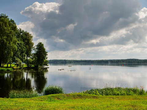 Lake scene with calm water reflecting cloudy sky. Trees line the shore, grassy bank in foreground. Distant boats float on still surface. Summer nature view scene. Calm and peaceful mood.