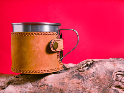 Stainless steel mug wrapped in brown leather sleeve. Mug rests on weathered wooden log. Vibrant red background behind. Leather strap with metal button closure