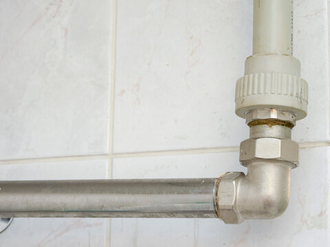White plastic pipe connects to metal elbow joint. Metal pipe runs horizontally against tiled wall. White tile wall has light gray grout lines. Scene shows plumbing installation detail