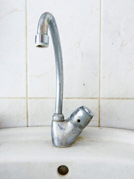 Old dirty metal faucet on white ceramic sink. Water tap shows signs of wear and rust. Background is tiled wall. Simple plumbing fixture close up shot. Suitable for renovation or retro-themed projects