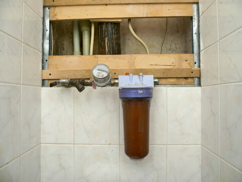 Water filter unit mounted on tiled wall. Pipes visible behind wooden frame. Brownish water inside filter housing. Metal fittings and pressure gauge attached. Typical home plumbing installation
