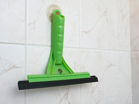 Green squeegee attached to wall via suction cup. Black rubber blade for window or glass cleaning. White tiled background with subtle gray veining. Tool hangs ready for use in bathroom or kitchen