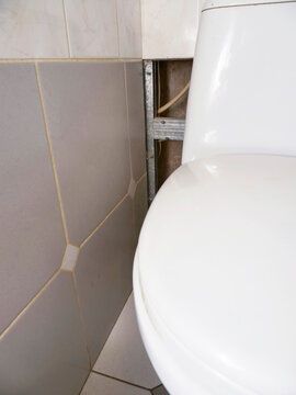 White toilet sits beside tiled wall. Wall has visible wooden framing and hole and needs repair work. Floor tiles match wall tiles. Image shows bathroom renovation process