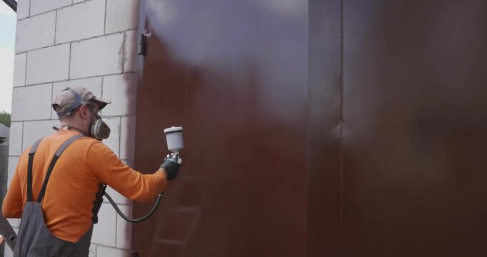 Professional worker in overalls and a respirator using an airbrush to spray paint a metal garage door brown, performing outdoor renovation and surface coating for corrosion protection.