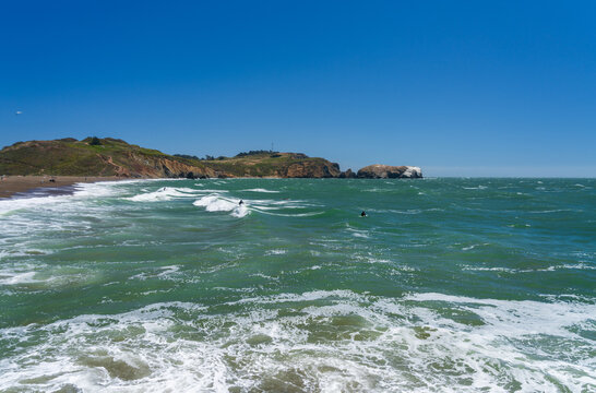 Surfers catch Pacific Ocean swells along the dark pebble shores of Rodeo Beach located within the scenic Marin Headlands of coastal California.