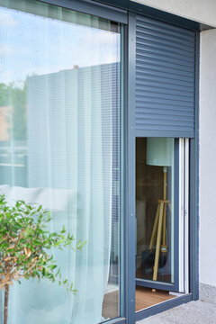 Roller shutter partially covers modern glass patio door beside sheer curtain and potted plant