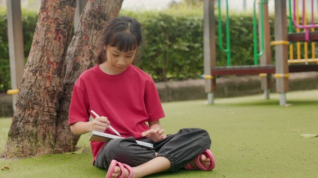 Asian kid sitting under a tree on the grass, drawing digital art on a tablet. Selective focus highlights the creative elementary student enjoying a relaxing outdoor hobby and personal time.