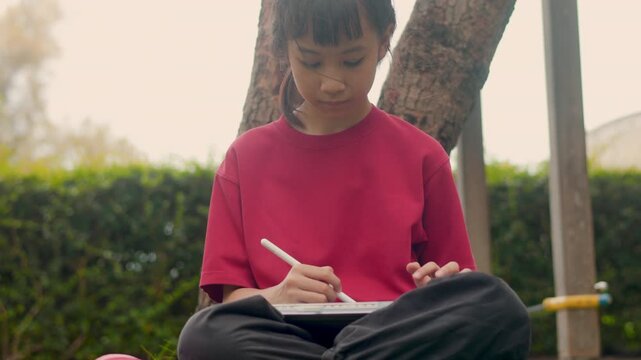 Selective focus captures the face of an Asian kid relaxing under a tree. With clear copy space, she enjoys drawing digital art and studying on a tablet during her casual outdoor hobby.