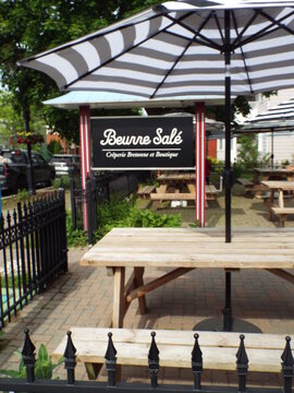 Gatineau, Quebec, Canada &ndash; June 7, 2026: Outdoor patio picnic tables with striped umbrellas stand in front of "Beurre Sal&eacute; cr&ecirc;perie bretonne et boutique" on Rue Principale.