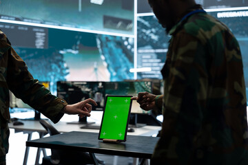 Obraz premium Army officers use a green screen tablet to review aerial surveillance imagery while military specialists analyze target data in a command center.