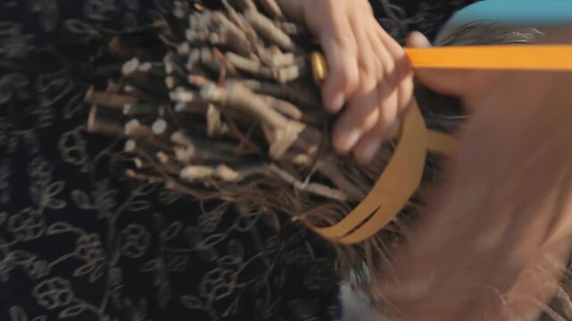 Close-up of hands tying dry twigs with a yellow strap to make a traditional handmade broom. Rural lifestyle, traditional crafting, and natural cleaning tool concept.