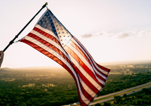 Large American Flag Blowing in the Wind From Tall Building Overlooking Scenic Green Landscape With Bright Sunset Sky