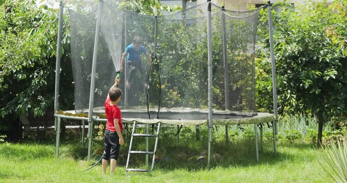 Kid spraying water from garden hose to his brother jumping in trampoline, summer fun
