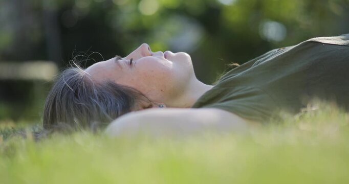 Mid-age woman laid back on green lawn and dreaming with closed eyes, day dreamer