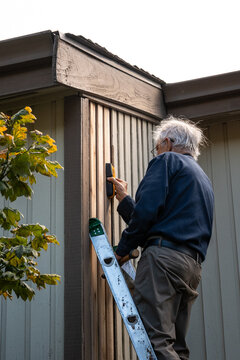 Elderly on an extension ladder using a stud finder installing new wood siding boards, homeowner maintenance and upkeep, making outdoor repairs on house
