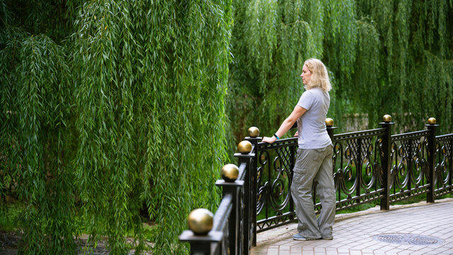 Middle-aged Caucasian woman standing by a metal fence in a park. Blonde female looking at lush green weeping willow trees. Casual outdoor lifestyle, background and leisure