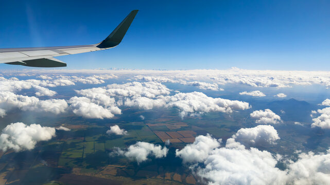 Aerial view from an airplane window showing fluffy clouds over patchwork agricultural fields. Commercial flight journey above the rural landscape. Plane wing in the blue sky