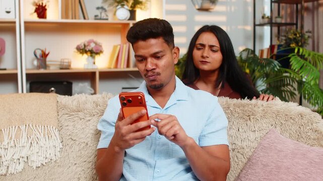 Indian couple man and woman sitting on home sofa read smartphone messages as woman peeks over shoulder and man notices hidden curiosity. Young family man and woman sitting on home sofa show surprise.