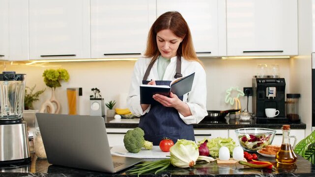Middle-aged woman smiling while taking diet notes on laptop in kitchen for balanced healthy eating routine. Young adult girl creating simple meal plan for weight loss using fresh food ideas at home.
