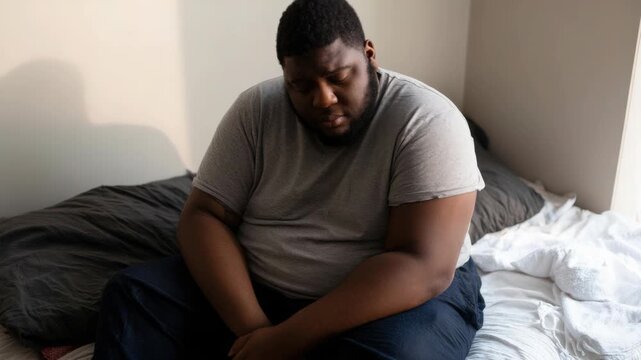 Depressed young African American man sitting alone on bed in bedroom, feeling sad and lonely, mental health concept, authentic candid video clip