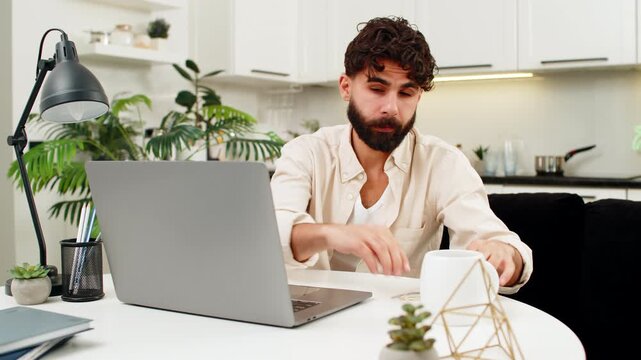 Indian man at home sits at laptop in glasses rubs tired eyes keeps typing text fights fatigue. Arabian guy sitting at table leans forward to screen works on project feels eye pain and stress inside.