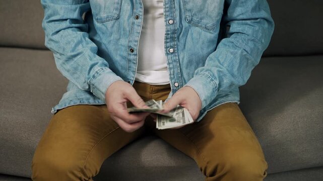 Close-up of a person counting a stack of US dollar banknotes while sitting on a sofa. Everyday money handling and personal finance concept focused on cash, savings, budgeting, income, finplanning.