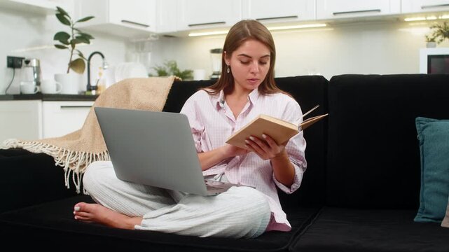 Young woman at home watches laptop lesson holds book and nods to guide for study and skills. Caucasian girl on sofa follows course video writes notes learns topics and keeps pace with focus and active