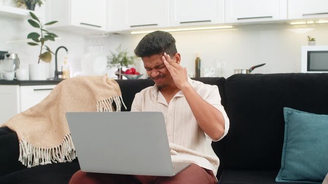 Indian man work on laptop at home rubs temples feels headache sharp pain. Arabian sick guy on sofa does short breathing drill closes eyes counts breaths for relief and resumes notebook tasks calmly