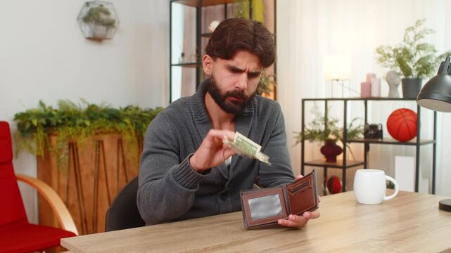 Indian man at home opens wallet finds only coins looks sad thinks about debt and overdue bills low pay. Arabian guy at desk plans budget counts money notes small income reads fees plans reduce spend