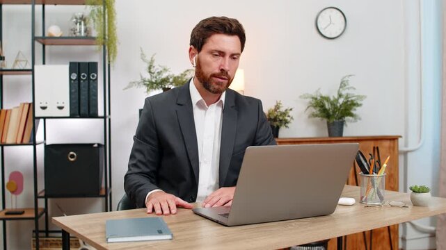 Middle-aged businessman at home office joins video conference on laptop wearing wireless earphones and waves hello. Freelancer guy at table smiles listens and takes notes in note book during meeting.