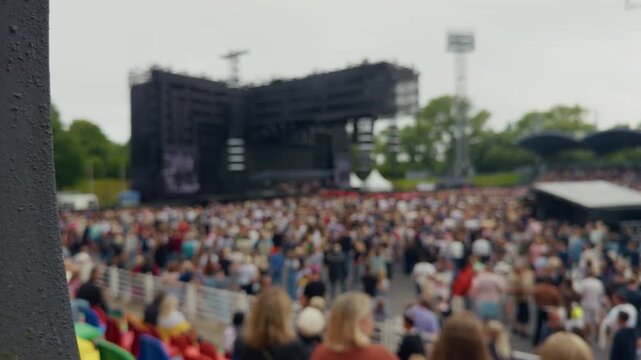 Large outdoor concert crowd gathered before blurred stage with foreground pillar and overcast sky at festival venue