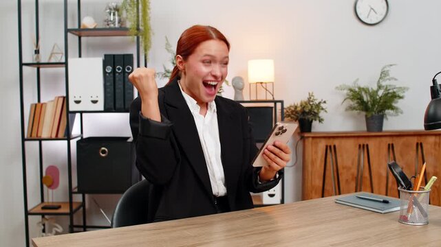 Young businesswoman at home office watches smartphone, bursts with joy after game win and positive good news Freelancer girl by table on chair pumps fist smiles celebrates victory and new opportunity.