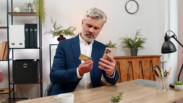 Middle-aged businessman at home office holds credit card taps smartphone confirming purchase enjoying easy checkout. Freelancer guy at table smiles relieved convenience saves time appreciates service.