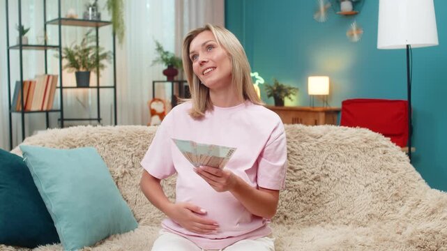 Pregnant middle-aged woman at home counting UAH Ukrainian hryvnia cash planning budget for childbirth banknotes serious focus. Girl reviews expenses money aiming safe future baby feeling responsible.