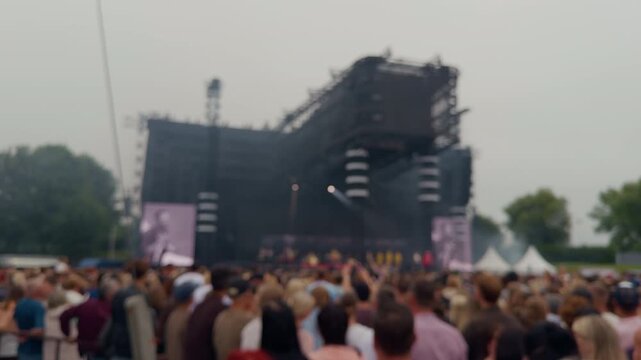 Blurred outdoor concert crowd facing large stage with screens and lighting tower under overcast sky