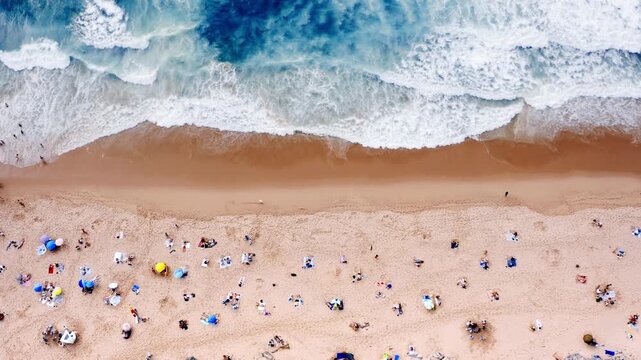 People relax on the beach under umbrellas aerial view. Some swim in the ocean while others walk along the shore. The sun shines brightly on this busy summer day.