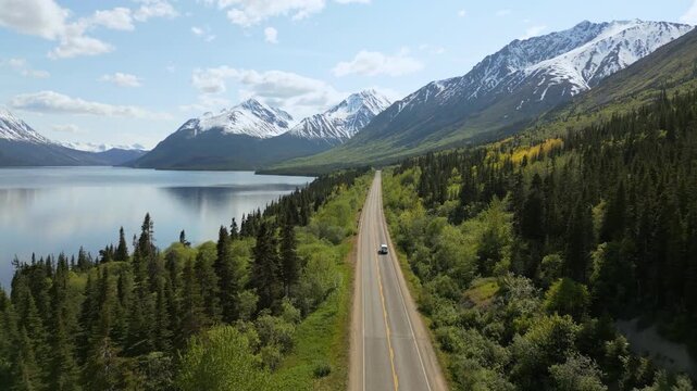 Drone aerial view of Camper van driving on a long road alongside a lake and snow capped mountains in Yukon, Canada