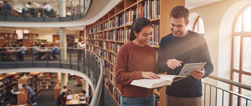 Students studying together in a modern library with warm sunlight rays, holding a book and digital tablet for university research, ideal for higher education e-learning concepts