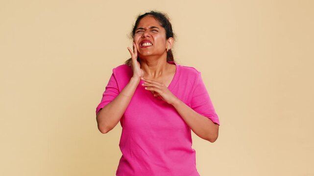 Indian woman touches cheek, visibly wincing in pain, clearly demonstrating severe toothache discomfort. Girl on beige background expresses dental pain discomfort distress urgently needing dental care