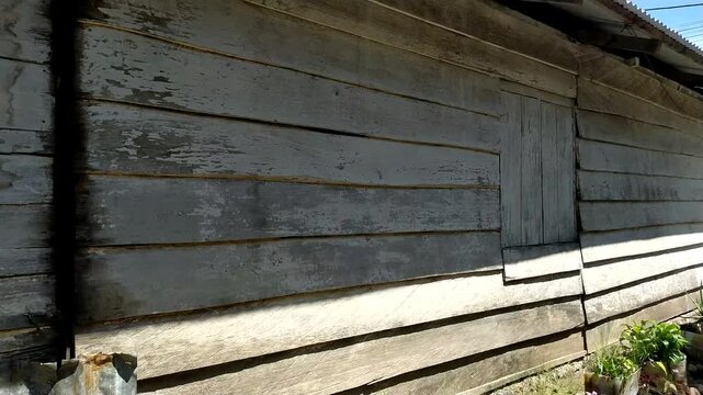 A side angle shot of an old weathered wooden wall with horizontal planks and a closed wooden shutter window of a traditional rural house, showing peeling white paint, aged timber texture