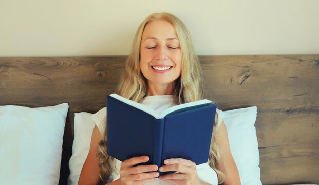 Happy middle aged woman reading book with blank cover while lying on the bed at home
